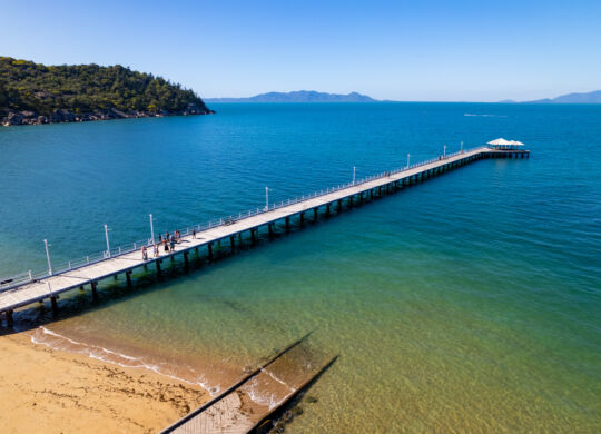 Drone shot of Picnic Bay Jetty. Empty beach in foreground, azure waters in the background